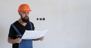 Mavi iş üniforması ve turuncu bir şapka giymiş, sakallı, vahşi bir inşaat işçisi. Üzerinde çizimler olan bir kağıt parçası tutuyor. Boşluğu kopyala.