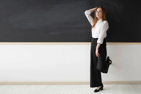 Stately beautiful young woman teacher at school. She is standing in front of a black blank board in her office. The girl is wearing a white shirt and black pants. She has youthful high-heeled shoes