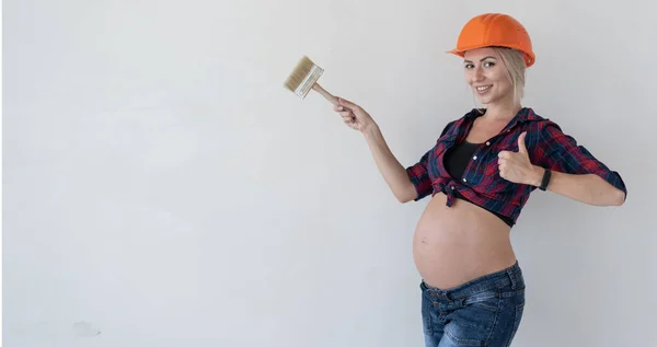 Молодая беременная женщина стоит на белом фоне. Одетый в рабочую одежду. Красная рубашка и строительный шлем. — стоковое фото