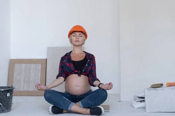 Blonde vrouw zit op de vloer in de kamer waar renovaties aan de gang zijn. over het hoofd wordt een beschermhelm gedragen. volledige Meditatie voor zwangere vrouwen. — Stockfoto