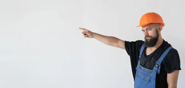 Un constructor en un casco naranja y uniforme está de pie en la habitación, señalando a un lugar vacío con su dedo. — Foto de Stock