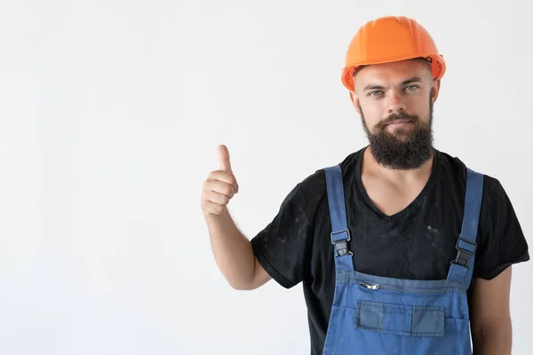 Un constructor con un casco y uniforme naranja está de pie en la habitación, levantando el dedo. mira al fotógrafo. — Foto de Stock