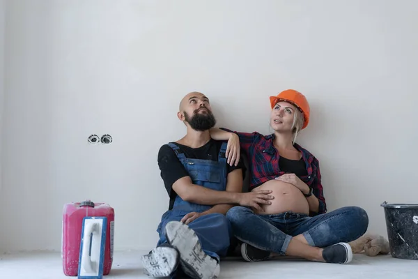 husband and his pregnant wife are sitting on the floor in the room. work uniform and orange safety helmet. stroking the belly and looking up at the empty space.