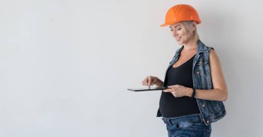 Kafasında turuncu koruyucu miğfer olan genç ve güzel bir kadın müteahhit. Hamile bir kız. Duvarın yanında elinde tabletle duruyor ve bir mesaj yazıyor. Doğum öncesi ev yenileme konsepti.