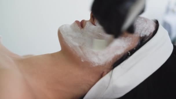 Kosmetikerin, die im Wellnessbereich ein enzymatisches Peeling auf das Gesicht von Frauen anwendet. Tragen Sie eine weiße Maske mit einem Pinsel auf. Kosmetischer Eingriff in einem modernen Schönheitssalon. — Stockvideo