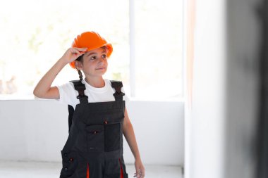 Kız tulum giymiş ve kafasında turuncu koruyucu bir kask var. 8 yaşında bir kız duvarın yanında duruyor ve ona bakıyor.