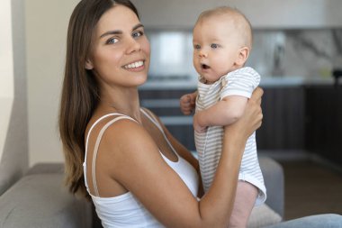 Genç ve güzel anne kanepede oturuyor. Yeni doğmuş bir bebeği kollarında tutuyor. Fotoğrafçıya bak.