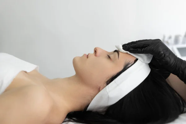 The beautician wipes the mask off the face of a young patient with cotton pads in the salon. Gloved hands of doctors wipe a woman's face. Cosmetic procedure in a modern beauty salon.
