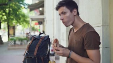 Genç beyaz erkek turist sokakta sosisli yiyor. Sırt çantası yakında. Yüksek kalite 4k görüntü