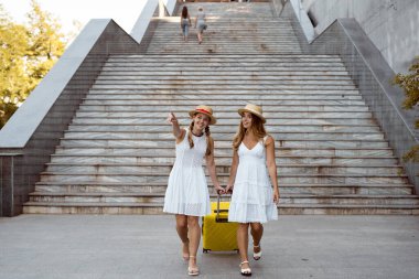 İki dişi ikiz yürüyüşçü merdivenlerin yanında yürüyor. Büyük sarı bir bavul çek. Metin için boş bir yeri parmakla göstermek.