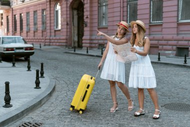 Bavullu gezginin genç kız arkadaşları otele gitmek için bir yol arıyorlar. Beyaz elbiseler ve güneş şapkaları giyiyorlar. Kızlar turizmi sever..