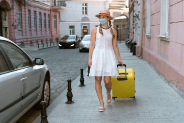 Koruyucu tıbbi maskeli genç bir kadın büyük sarı bir bavulla caddede yürüyor..