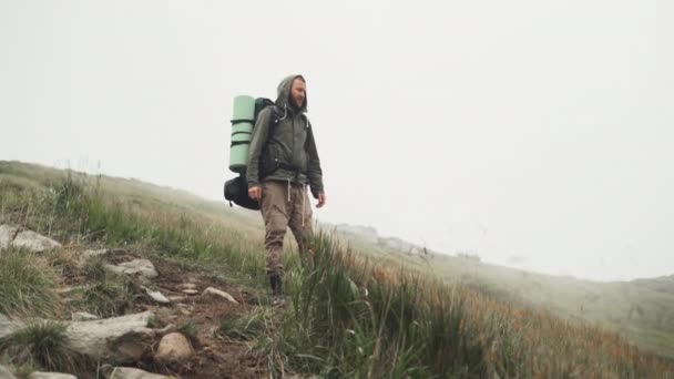 Een man toerist met een grote rugzak staat op de top van de heuvel en bewondert het landschap — Stockvideo