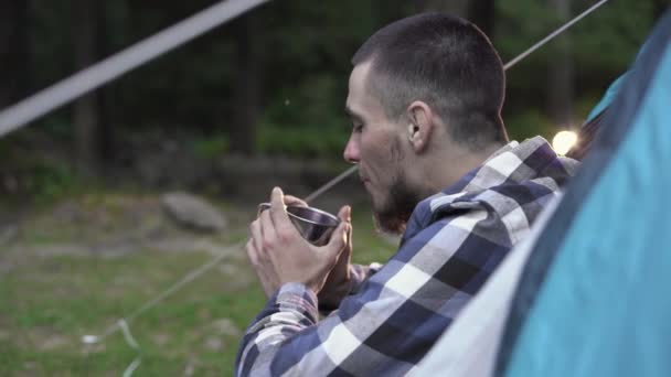 Jovem senta-se em uma tenda e bebe uma bebida quente de um copo. — Vídeo de Stock