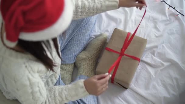 Vista superior de una chica en un suéter y un sombrero de Navidad rojo se sienta en la cama y abre una caja con un regalo — Vídeo de stock