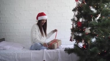 Süveter ve kırmızı Noel şapkalı bir kız yatağa oturur ve hediyeli bir kutuyu açar.