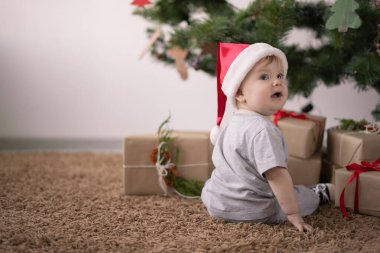 Noel Baba şapkalı küçük bir çocuk Noel ağacının yanında yerde oturuyor ve arkasına bakıyor..