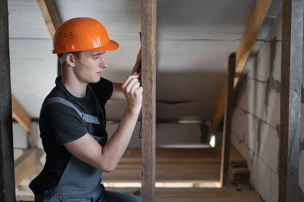 male builder dressed in work clothes and an orange hard hat. Measures the distance with a tape measure and puts a mark on the board. Copy space