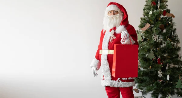 サンタクロースは片手に大きな買い物袋を持ってクリスマスツリーの近くに立っています バナー コピースペース — ストック写真