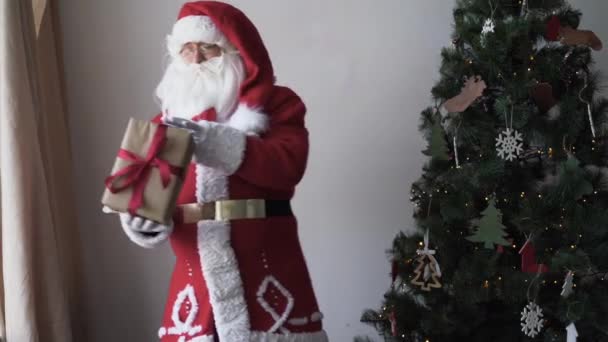 Retrato do feliz Papai Noel. Fica perto da árvore de Natal segurando uma caixa com um presente — Vídeo de Stock