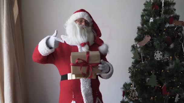 Potret bahagia santa claus. Berdiri di dekat pohon Natal memegang kotak dengan hadiah dengan jari telunjuk menunjuk ke sana — Stok Video