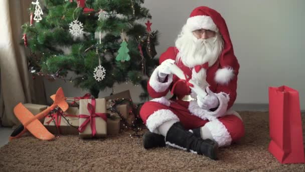 Père Noël est assis sur le sol près de l'arbre de Noël, tenant une feuille de papier roulé dans ses mains — Video