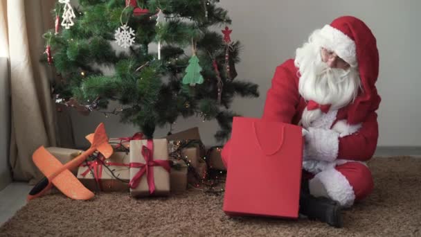 サンタクロースはクリスマスツリーの近くの床に座って、大きなショッピングバッグから雪の結晶を取り、それを飾る. — ストック動画
