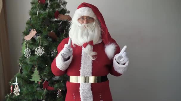 Santa claus se tient près du sapin de Noël pointe avec ses doigts à un espace vide pour le texte — Video
