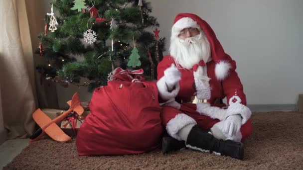 Santa Claus sedí u vánočního stromku a s někým komunikuje — Stock video