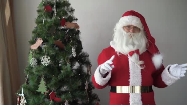 Santa Claus se encuentra cerca del árbol de navidad señala con sus dedos a un espacio vacío para el texto — Vídeos de Stock