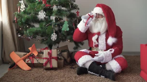 Santa claus sits near a christmas tree looks at a rolled up scroll and is surprised at what he sees — Stock Video