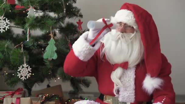 Santa claus duduk di dekat pohon natal melihat gulungan gulungan yang digulung dan terkejut dengan apa yang dilihatnya — Stok Video