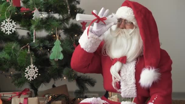 Santa Claus sedí na podlaze poblíž vánočního stromku, drží v rukou srolovaný list papíru, dívá se skrz něj jako dalekohledem — Stock video