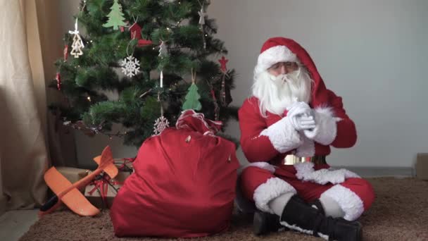 Santa claus sits on the floor near the christmas tree, unties the bag and puts his hand in it — Stock Video