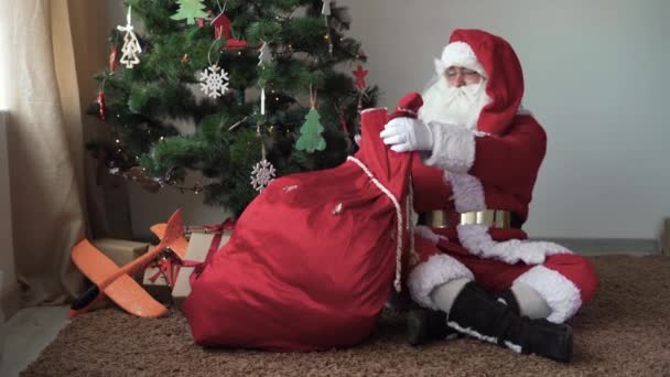 सांता क्लॉस भेटवस्तू एक पिशवी बांधून ख्रिसमस ट्री जवळ मजलावर बसतो — स्टॉक व्हिडिओ