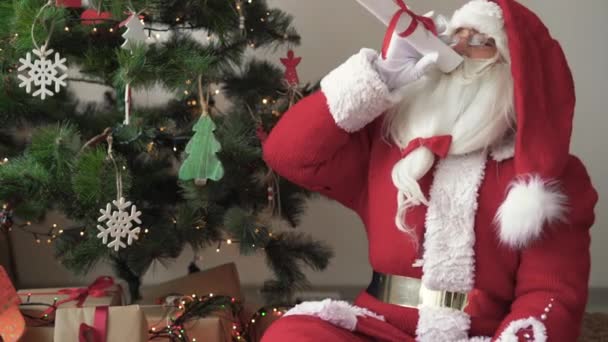 Santa claus sits near the christmas tree takes a rolled-up roll of paper and shouts into it like a loudspeaker — Stock Video