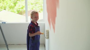 Avrupalı görünümlü çocuk evin duvarını bir silindirle boyuyor. Tulum giymişti.