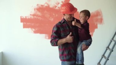 Kollarında bir çocuk tutan bir adam, kafasına koruyucu bir kask takıyor. Başparmak yukarı.