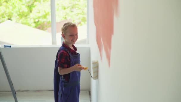 Avrupalı görünümlü çocuk evin duvarını bir silindirle boyuyor. Tulum giymişti. — Stok video