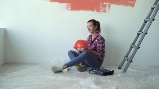 En kvinna sitter på golvet i ett hus, sätter en orange skyddshjälm på huvudet. Renoveringskoncept för hemmet — Stockvideo