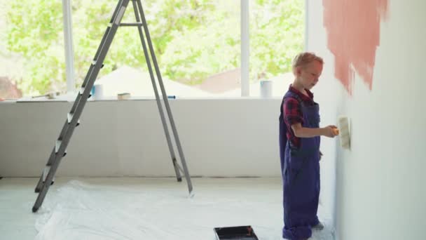 Chłopiec o europejskim wyglądzie maluje ścianę domu rolką. Koncepcja pomocy rodzicom — Wideo stockowe