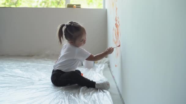 Маленькая девочка сидит на полу возле стены и рисует на ней оранжевой краской — стоковое видео