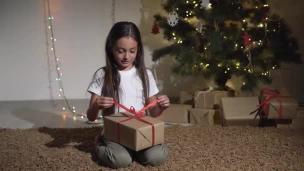 Маленькая девочка сидит рядом с елкой, развязывает бант на коробке с подарком — стоковое видео
