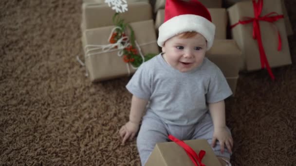 Little child in santa claus hat sits on the carpet. Many boxes with gifts — Stock Video