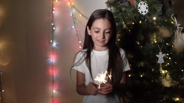 Ένα μικρό κορίτσι στέκεται κοντά σε ένα χριστουγεννιάτικο δέντρο κρατώντας φλεγόμενες σπίθες στα χέρια της — Αρχείο Βίντεο