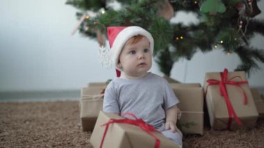 Noel Baba şapkalı küçük çocuk içinde hediye olan bir kutu tutuyor. Noel ağacının yanında yerde oturuyor.