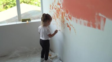 Küçük bir kız duvarın yanında duruyor ve bir boya fırçasıyla resim çiziyor. Turuncu boya