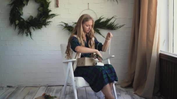 Jovem mulher bonita está sentada em uma poltrona e embalando um presente. — Vídeo de Stock