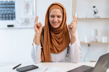Umut dolu Müslüman kadın doktor parmaklarını çapraz yapıp gülümsüyor, dilek diliyor ve hastane ofisinde dua ediyor. Boşluğu kopyala