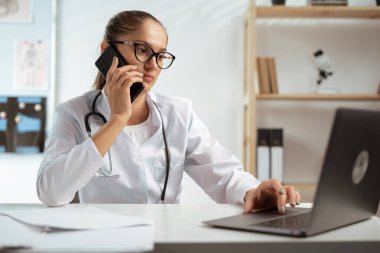 Genç doktor kadın cep telefonu danışmanlığı yapan bir hastayla konuşuyor, iş yerinde dizüstü bilgisayar kullanıyor. İletişim için tıp ve teknoloji kavramı